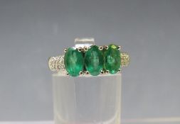 AN 18CT WHITE GOLD 'ILIANA' THREE STONE EMERALD RING, with diamond chip set shoulders, ring size O t