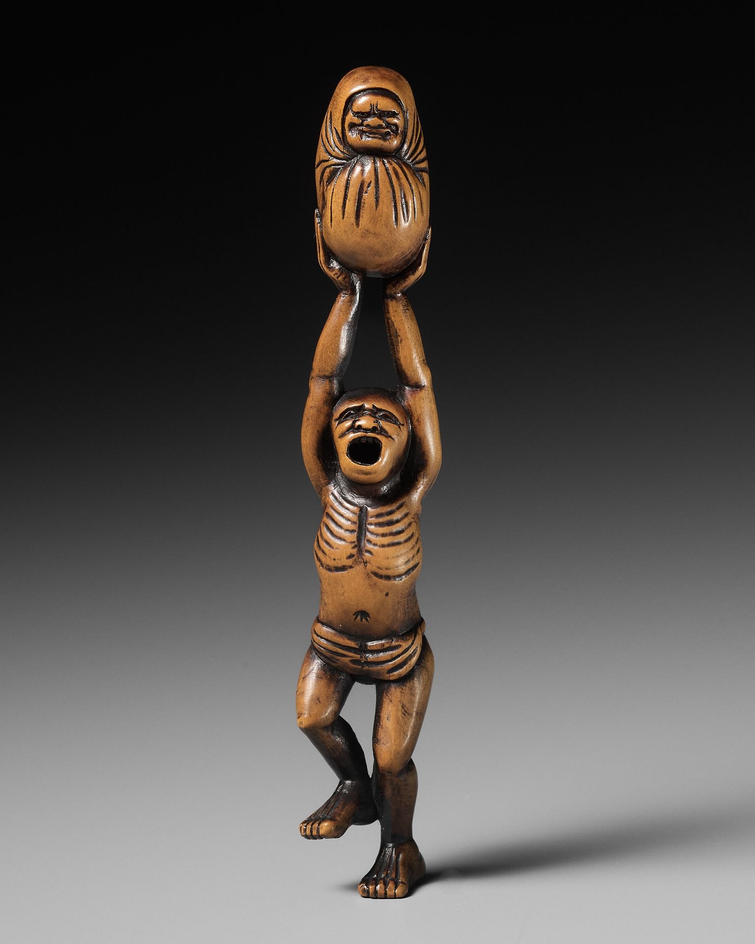 A POWERFUL WOOD NETSUKE OF A DANCING MAN WITH A DARUMA DOLL