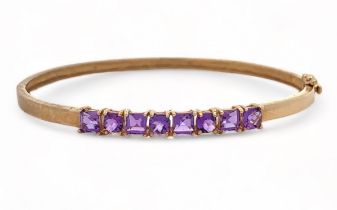 A small 9ct gold hinged bangle set with eight purple gemstones, likely CZs, alternating square cut