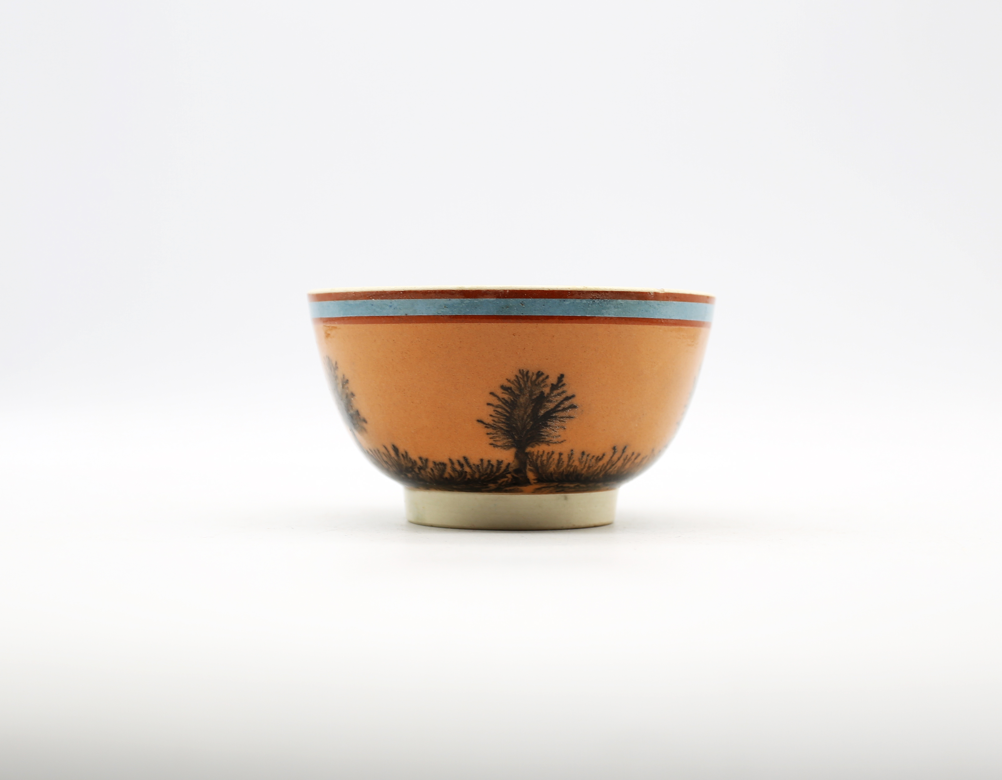 A small creamware Mocha bowl, orange ground, with black trees and a blue band.  Circa 1800-1820.