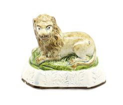A pearlware model of a recumbent lion laying on a grassy mound on a base with moulded relief.  Circa