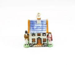 A small Prattware money box in the shape of a house, flanked with a man and woman on either side.