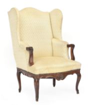 An 18th century wingback armchair, with carved walnut open arms, shaped apron with carved central