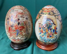 Two Satsuma-style porcelain eggs, both hand painted with Oriental Geisha scenes on wooden bases,