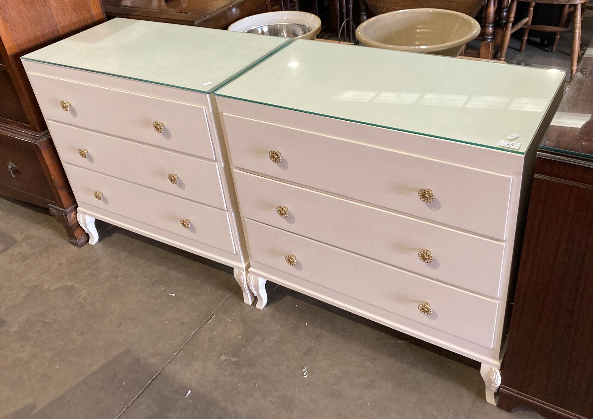 A pair of cream painted wooden three-drawer bedside chest of drawers with glass protective tops,