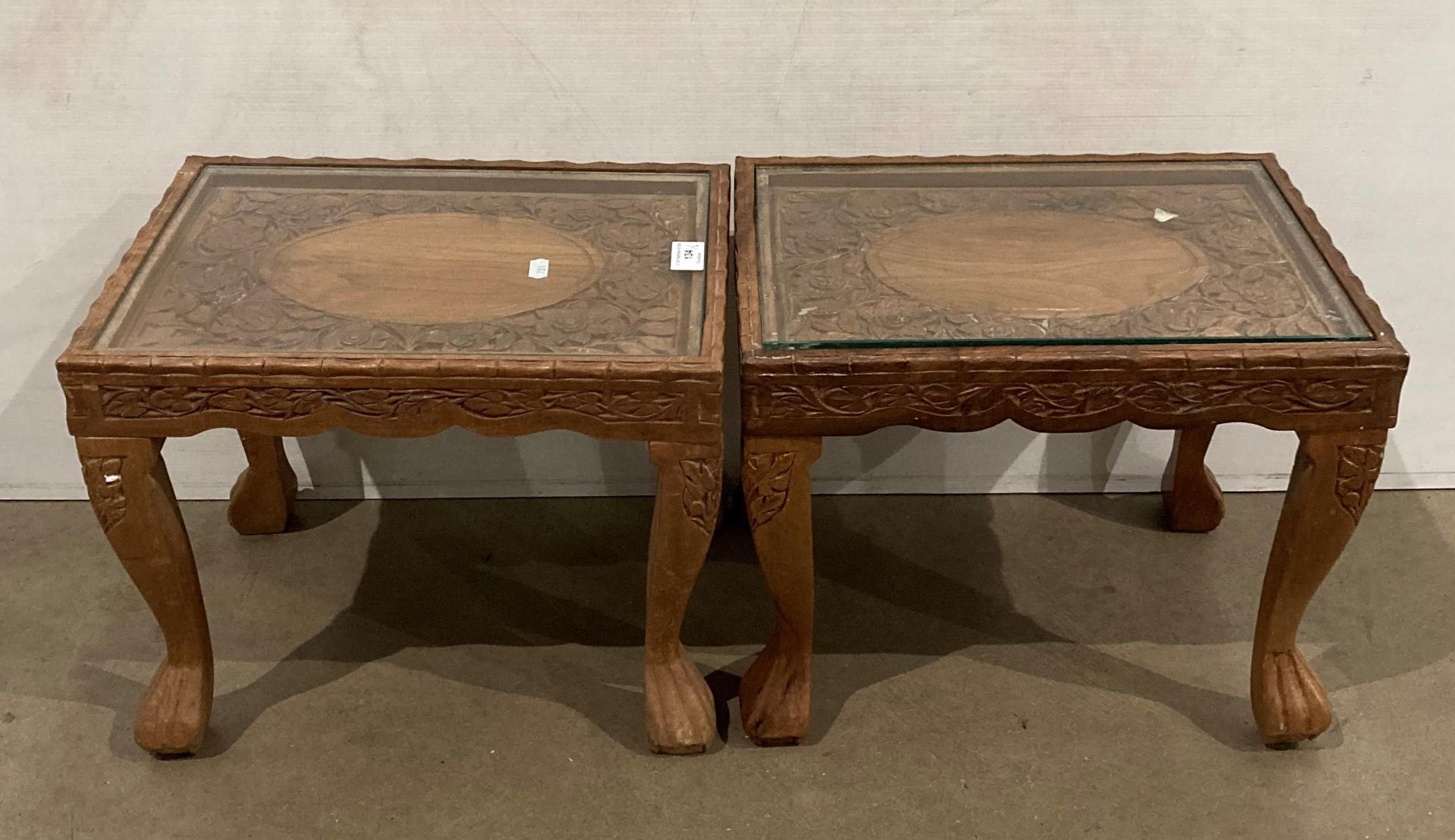 A pair of Asian hand-carved wooden tables with glass tops and with a floral design to each table,