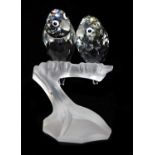 A pair of Swarovski crystal parakeets, modelled perched on the frosted glass branch of a tree stump,