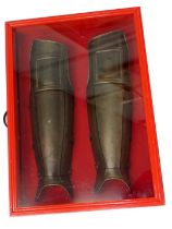 Two replica armour leg guards, in red painted and perspex case, 75cm wide.