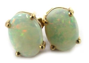 A pair of opal stud earrings, each set with an oval opal in four claw basket setting, on a yellow me