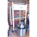 A large steel milk pail, large cooking vessel made of aluminium and a two tier trolley with drawer.