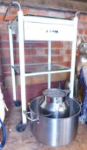 A large steel milk pail, large cooking vessel made of aluminium and a two tier trolley with drawer.