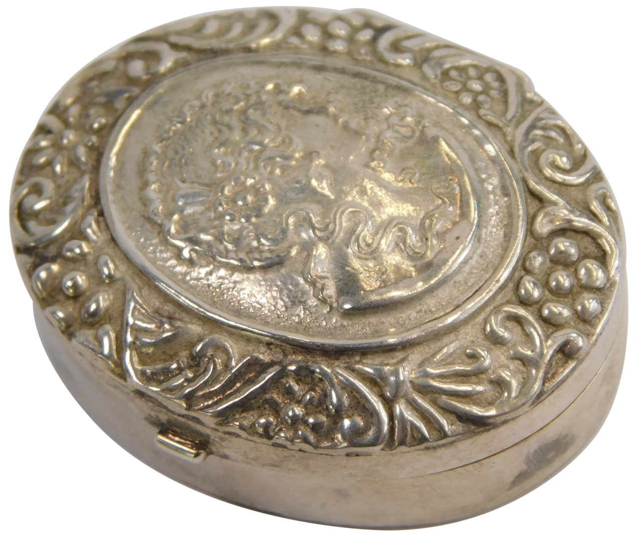 A white metal pill box, of circular design with hammered female head and flowing scroll floral
