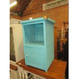 A turquoise painted basket weave bedside cupboard, with a glass top, shelf and single door.