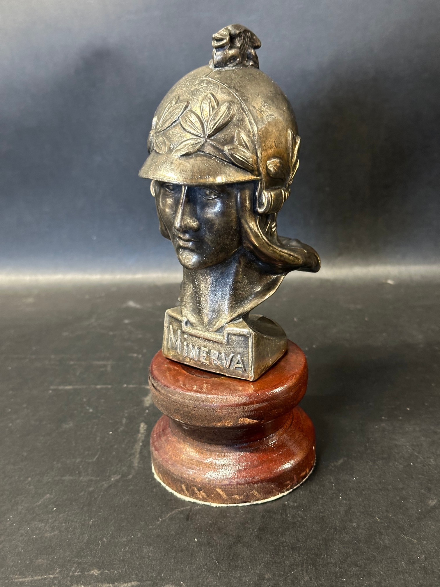 A Minerva (Roman Goddess) car accessory mascot with griffin on helmet, mounted to wooden base.