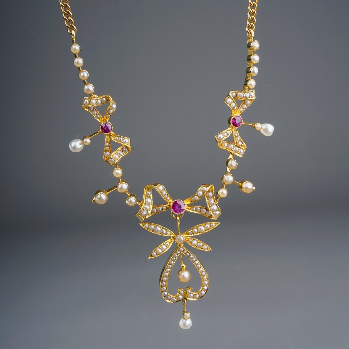 An Edwardian yellow gold ruby and seed pearl necklace, three pearl-set bow motifs each centered with