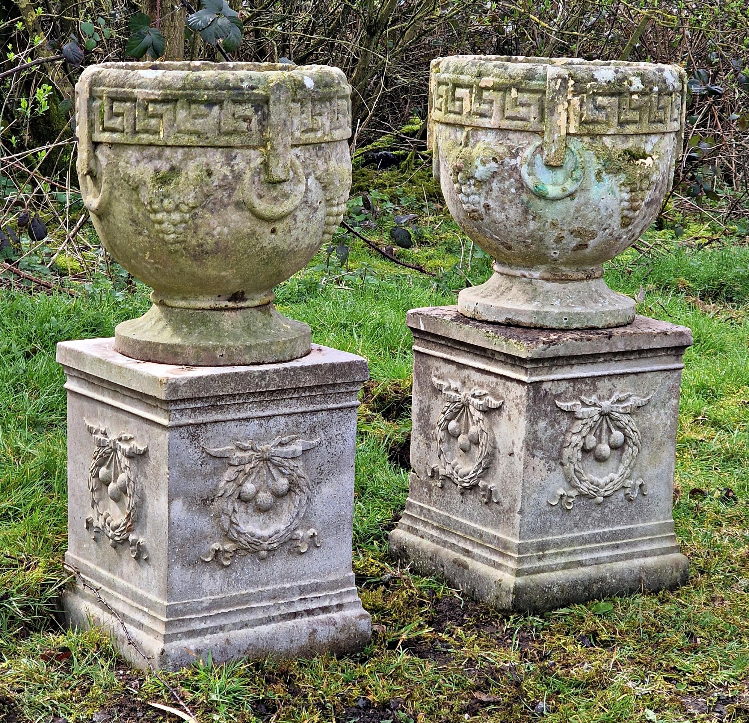 A pair of weathered reconstituted stone garden urns with Greek Key and grape detail raised on