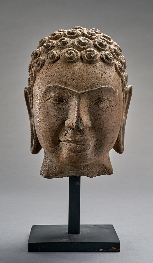 Kopf eines Buddha. Betonte Brauenpartie. Lächelnd. Frisur mit geschneckten Haarlocken und ungewöhn