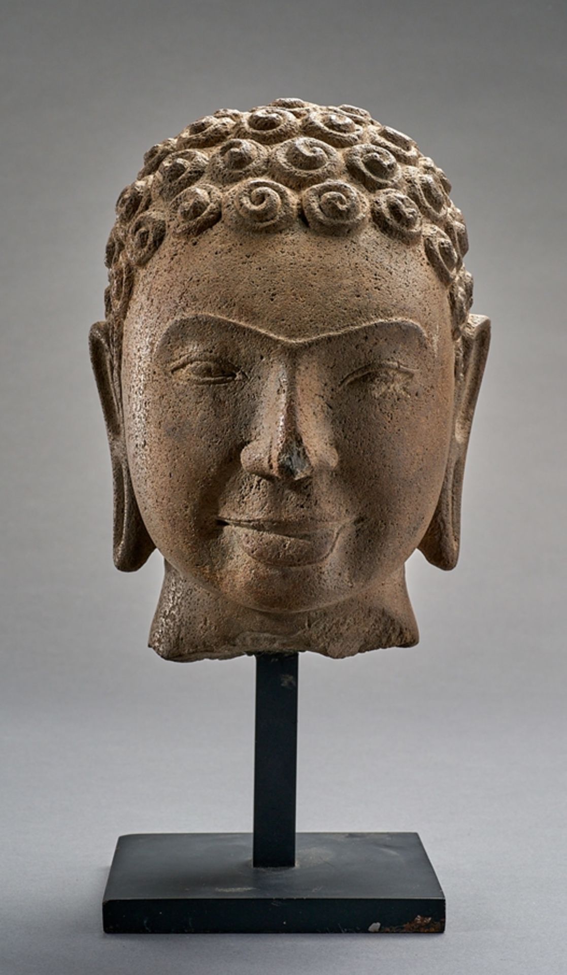 Kopf eines Buddha.  Betonte Brauenpartie. Lächelnd. Frisur mit geschneckten Haarlocken und ungewöhn