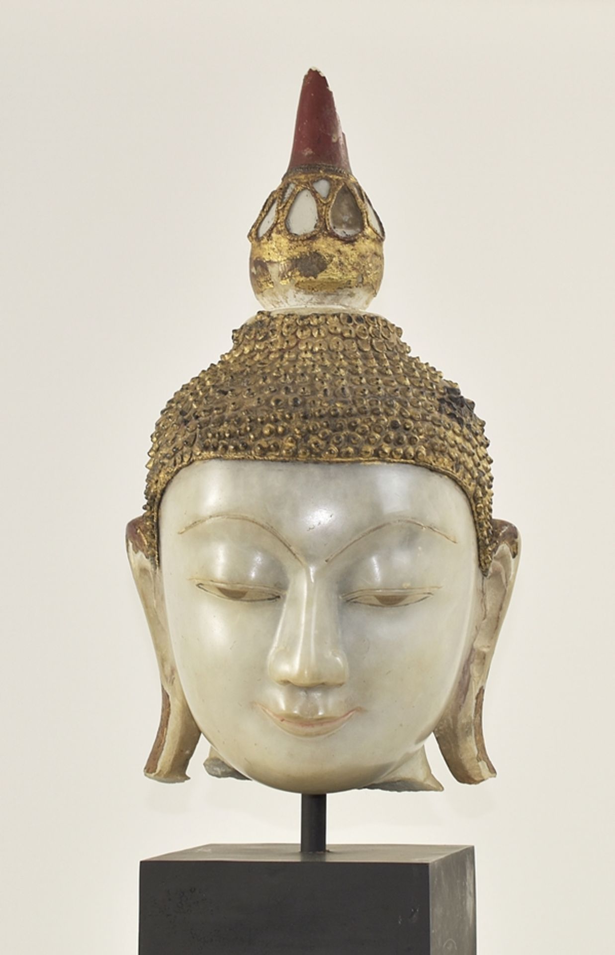 Kopf eines Buddha. Lächelnd mit gesenkten Augenlidern. Lange Ohren. Frisur mit kleinen Locken. Verz