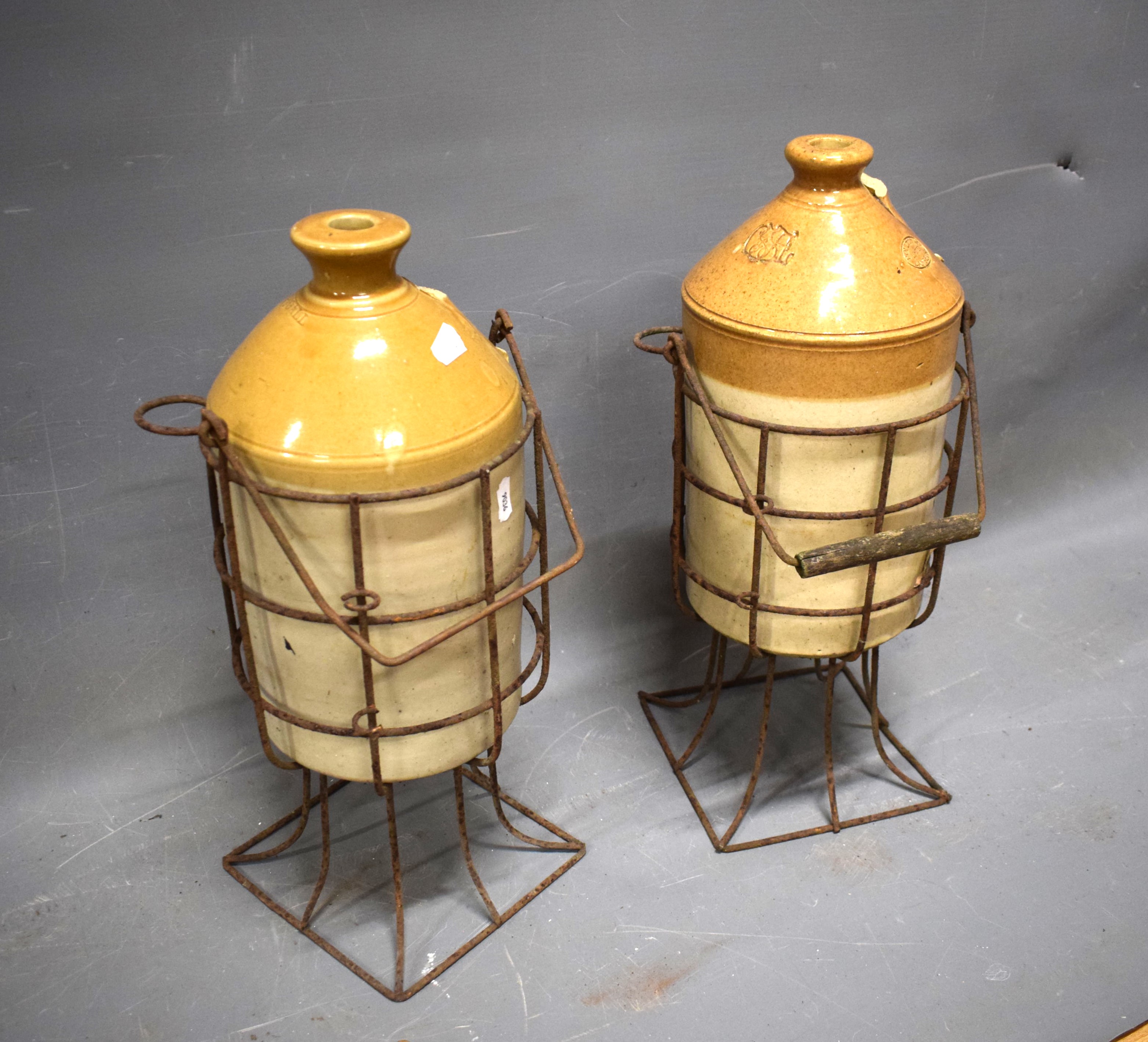 Two Stoneware flagon jars cradled inside a wire crate. Both jar handles missing. Each approx 19 inch