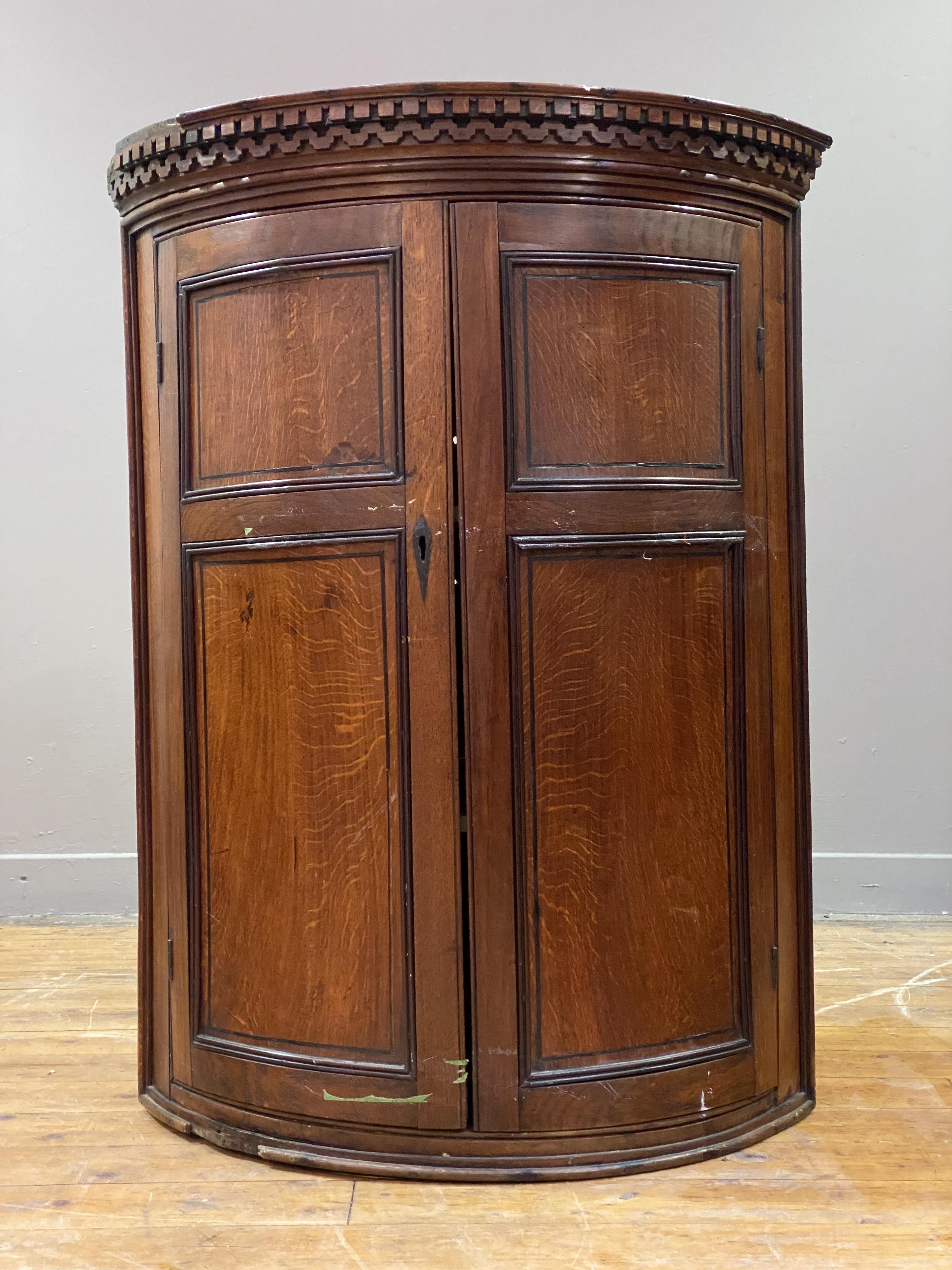 A Georgian oak bowfront wall-hanging corner cupboard, the dentil cornice above two twin-panelled