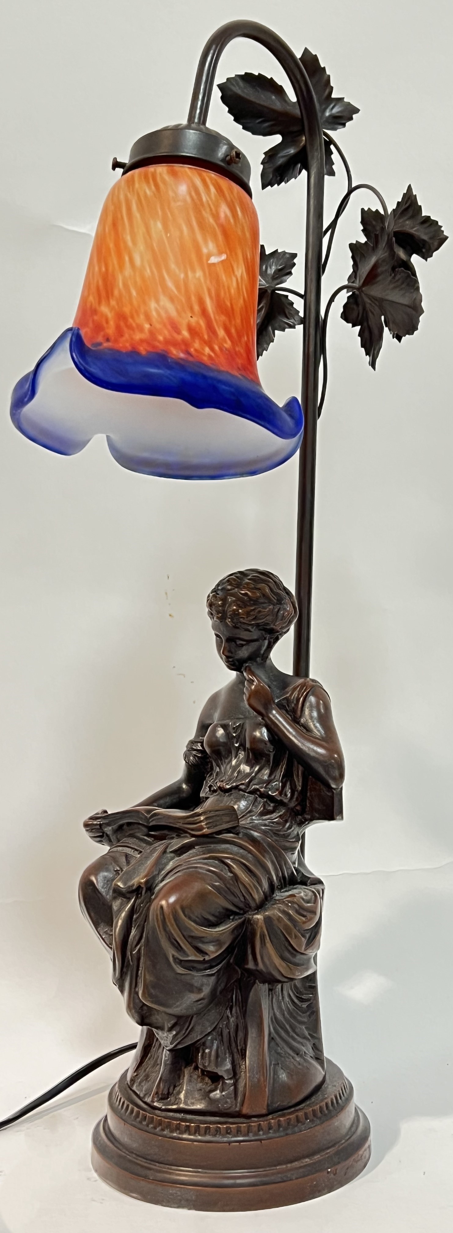 A bronze tone resin figural lamp modelled as a lady reading with trefoil coloured glass shade and
