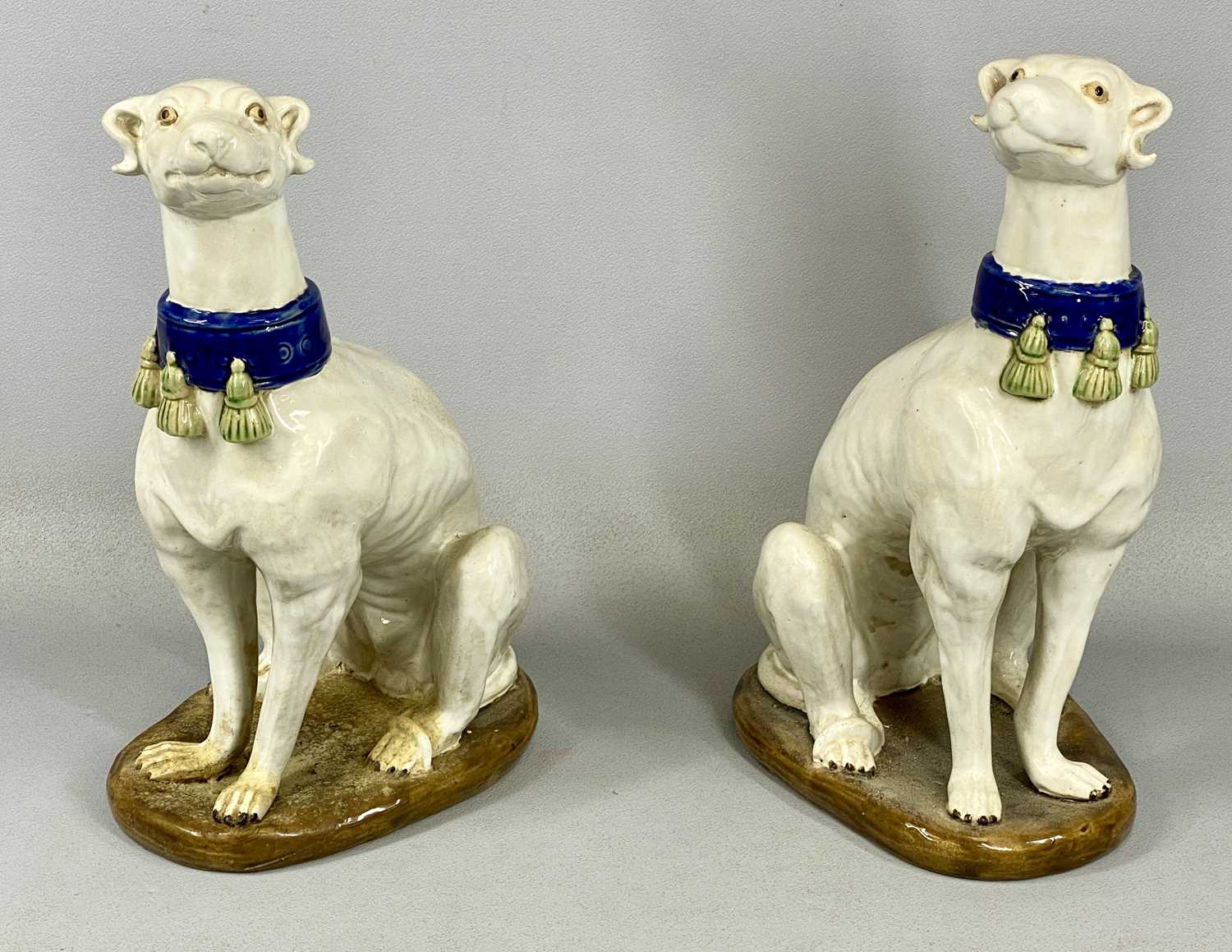 PAIR OF ITALIAN MAJOLICA GREYHOUNDS 20th century, seated and in cream glaze with tassled blue