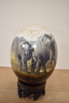 A 20th Century African design ostrich egg, decorated with elephants, and raised on a carved hardwood