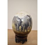 A 20th Century African design ostrich egg, decorated with elephants, and raised on a carved hardwood