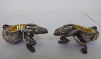 A pair of parcel gilt and white metal novelty salt cellars, each fashioned as a frog, pulling a