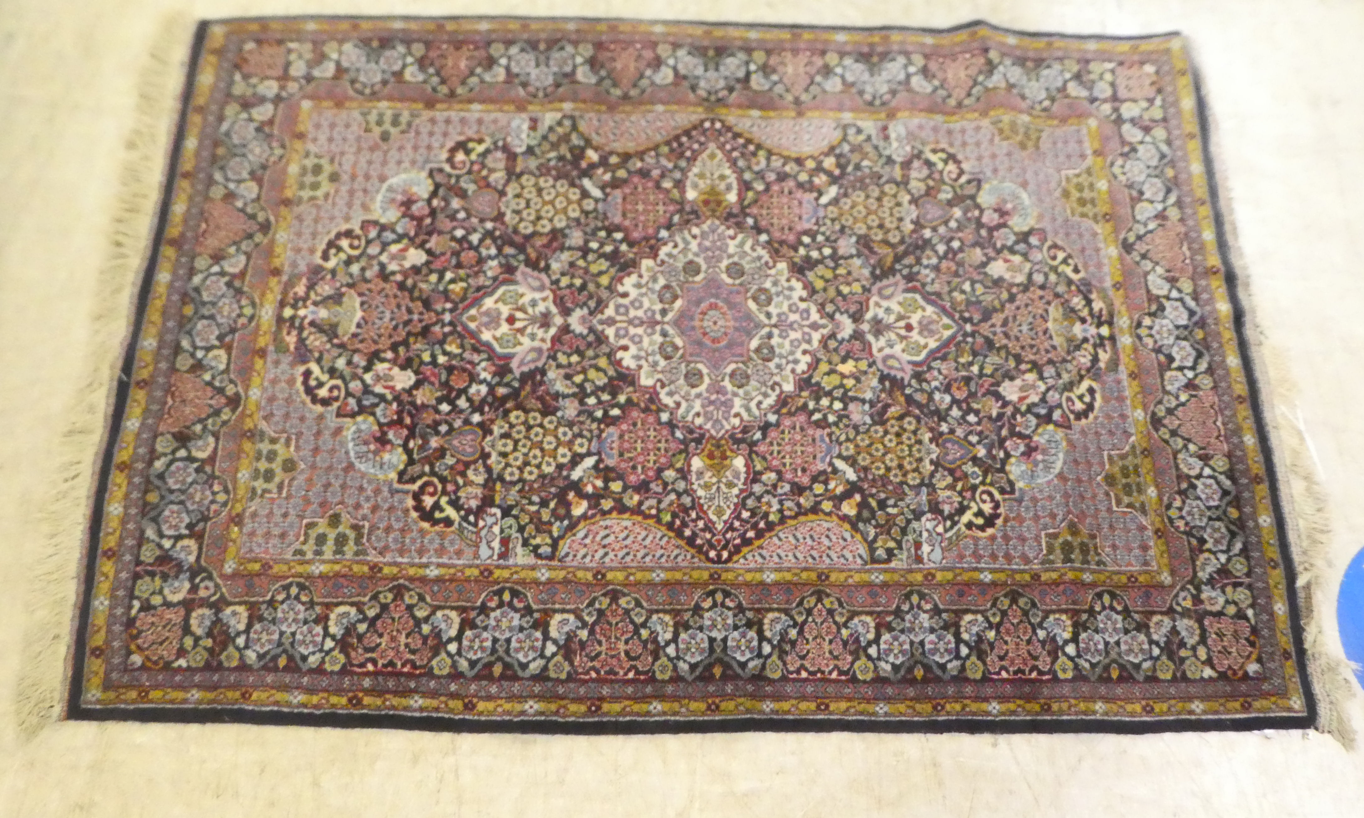 A Persian rug, decorated with a central motif, bordered by stylised designs, on a multi-coloured