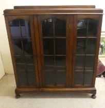 A 1930s oak display cabinet with three partially glazed doors, on a plinth and bun feet  49"h  45"w