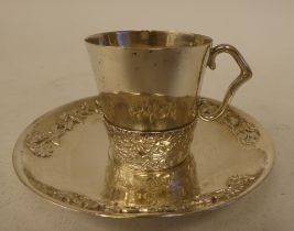 An Oriental silver coloured metal coffee cup and saucer, decorated in cast and applied floral