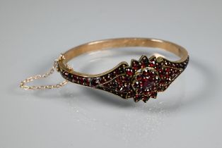 A Victorian garnet-set bangle, the oval half-hinged yellow metal bangle with central flower motif,
