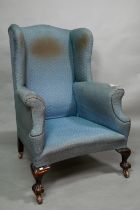 A traditionally upholstered Queen Anne style wing armchair, with concave seat front, raised on short