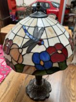 A Tiffany style lamp with a glass shade applied with a dragonfly and flowers on a naturalistic base