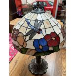 A Tiffany style lamp with a glass shade applied with a dragonfly and flowers on a naturalistic base