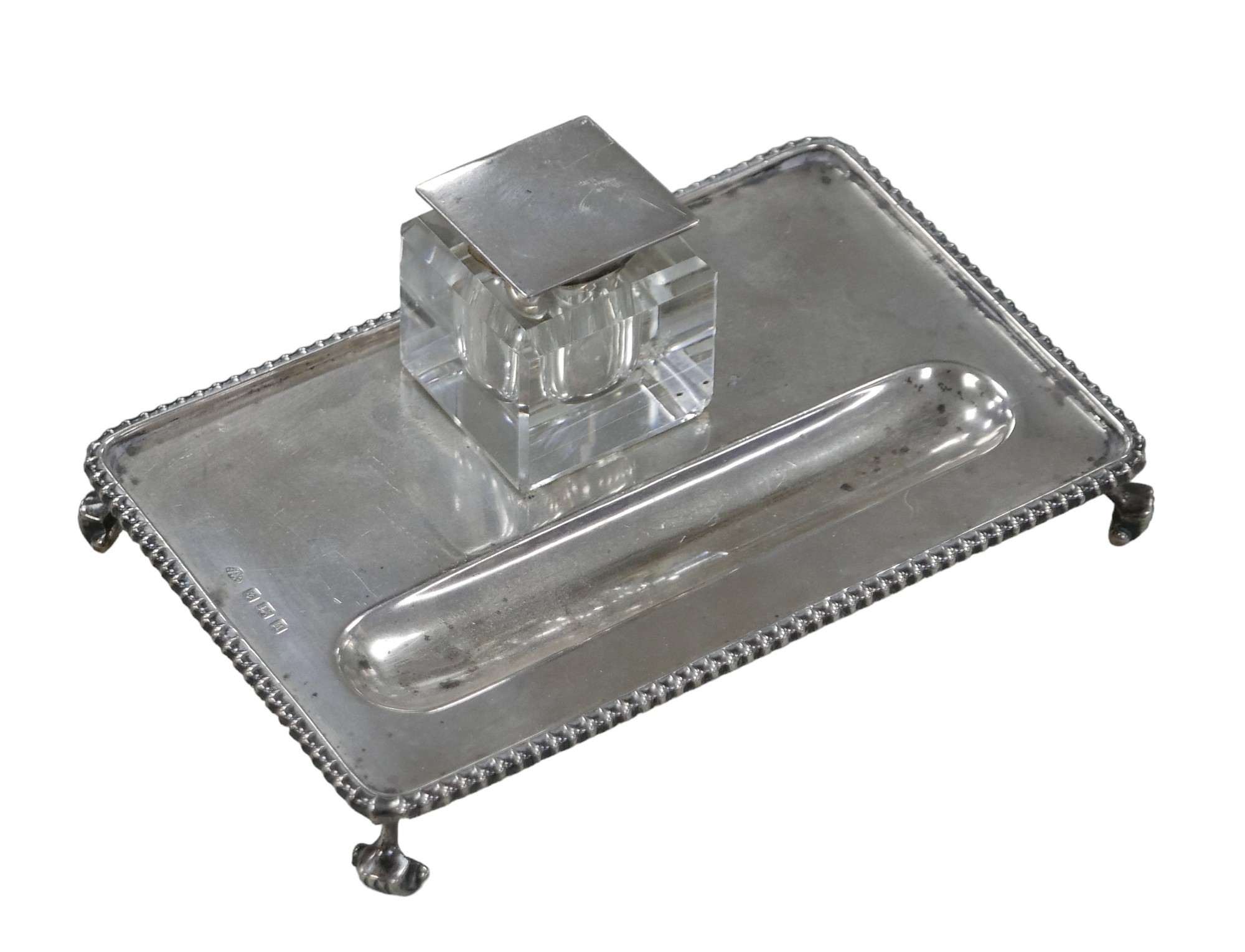 A George V silver inkwell stand raised upon four scroll feet, with silver topped glass inkwell, Adie