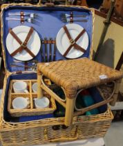A wicker picnic basket with contents with a wicker stool