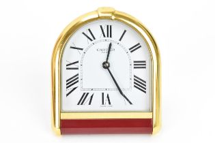 A Cartier travel alarm clock, having an arched top gilt metal case with red lacquer below, white