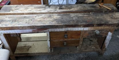 A wooden workbench with an iron vice to the right side, drawers and open storage spaces, on block