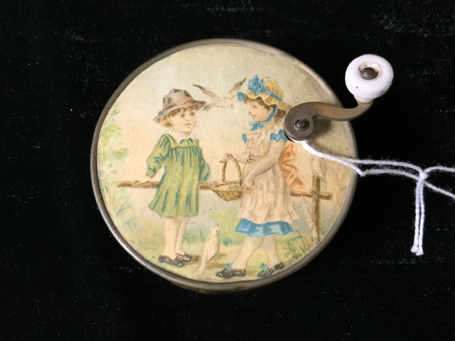 A VINTAGE CIRCULAR MUSIC BOX WITH TOP HANDLE DECORATED WITH A CHILDHOOD SCENE TO TOP; EMBOSSED HARPS - Image 2 of 3