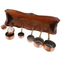A graduated set of copper pans with brass handles, largest 10cm diameter, with an oak wall-mounted