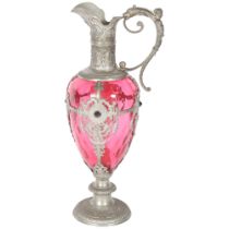 A cranberry glass Claret jug, with ornate allover pewter mounts and scrolled handle (no stopper),