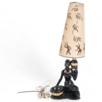 A painted plaster table lamp in the form of a Nubian style female figure, with associated shade,