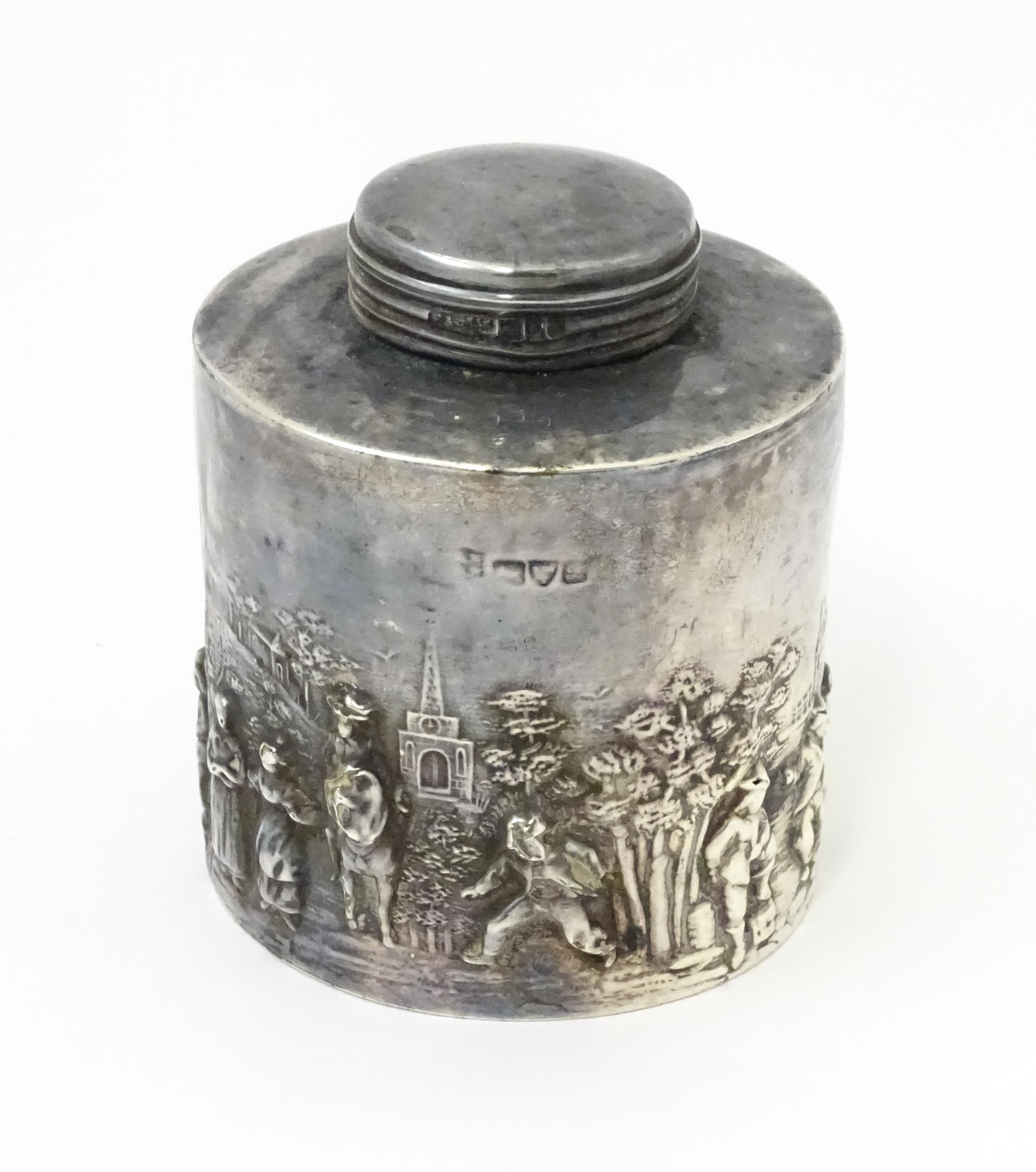 A silver tea caddy depicting a village scene with buildings and figures. Hallmarked Chester c.