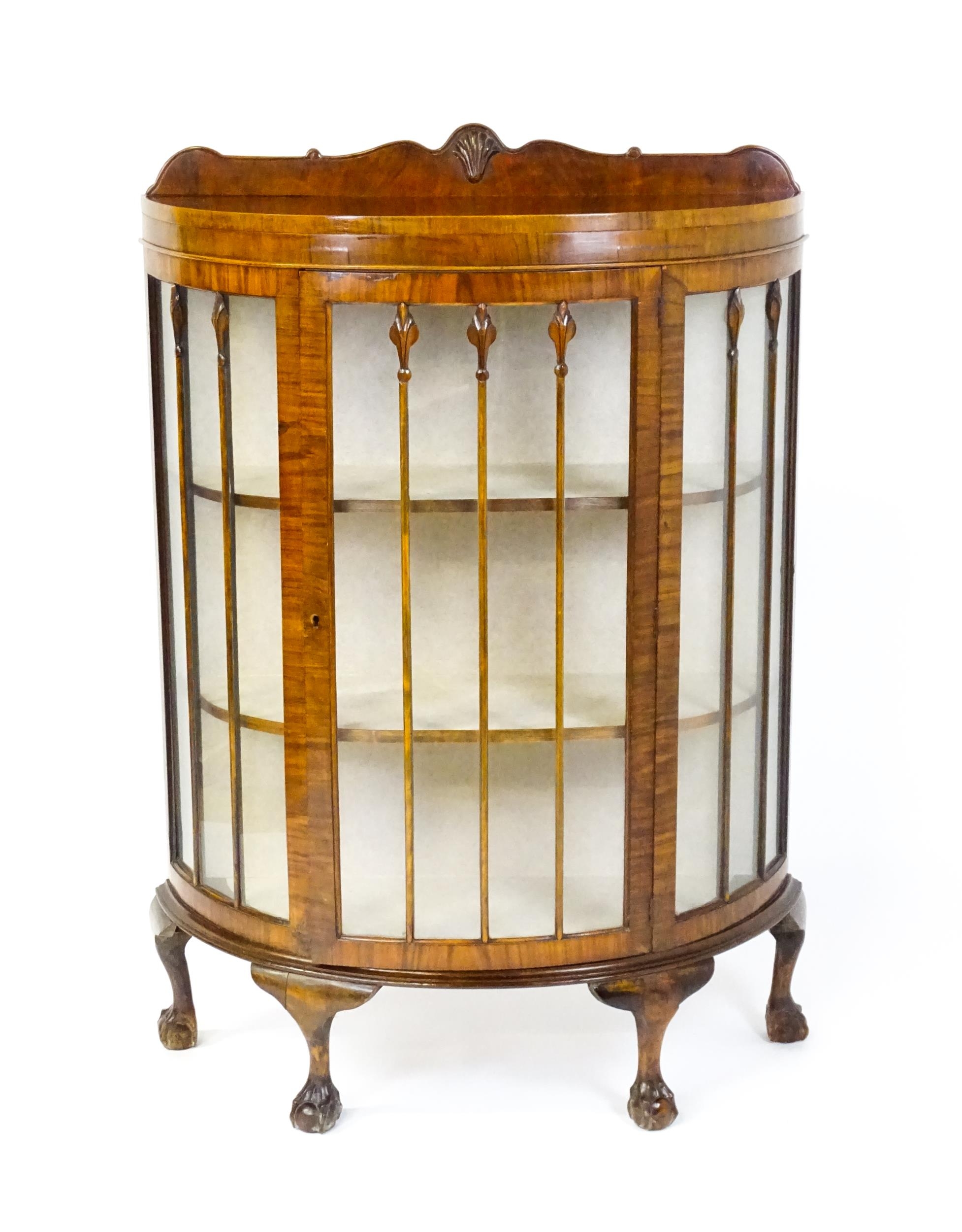 An early / mid 20thC walnut demi lune display cabinet with a shaped and carved upstand above a