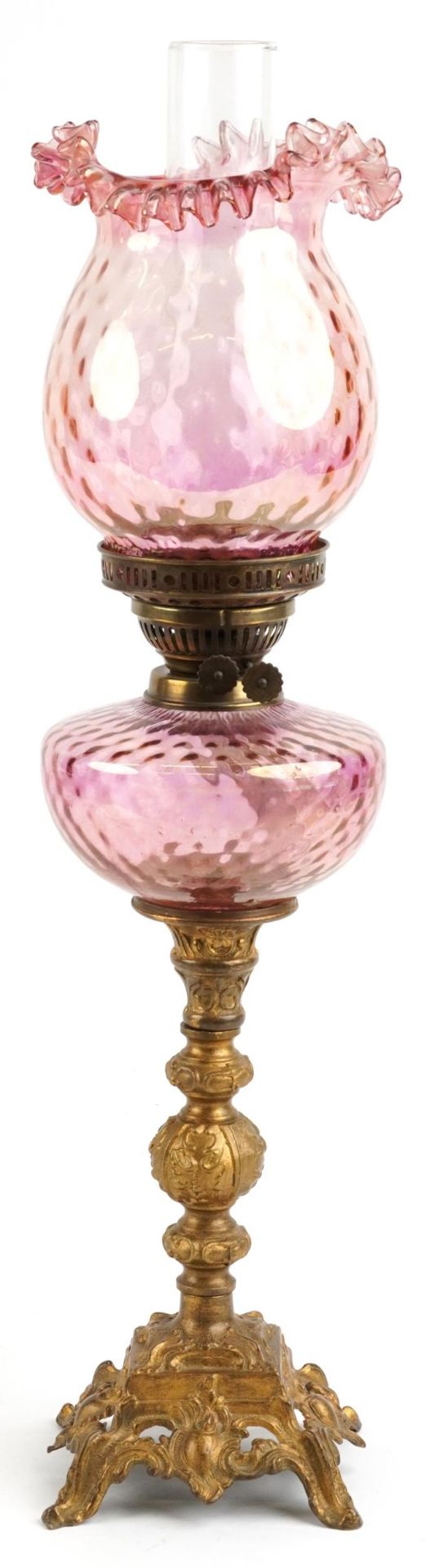 Ornate gilt painted oil lamp with iridescent cranberry glass shade and reservoir, 68.5cm high :