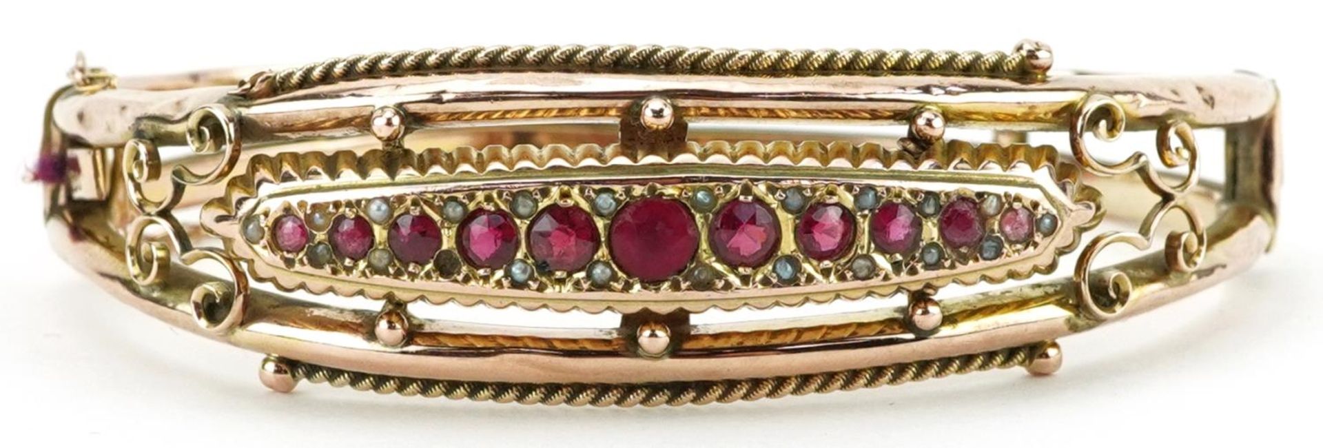 Edwardian 9ct rose gold hinged bangle set with eleven graduated red stones and seed pearls,