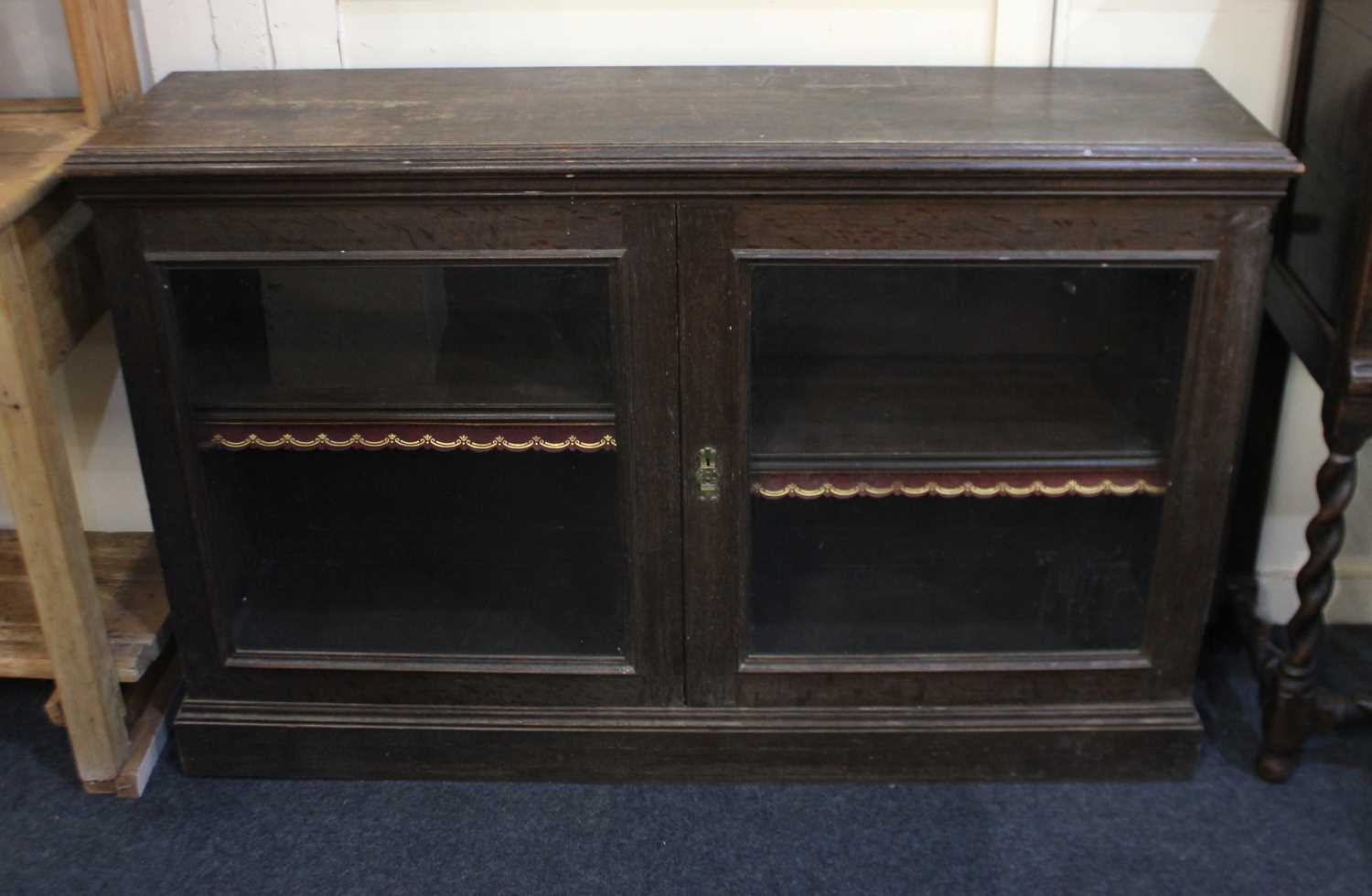 An oak cabinet with two glazed panel doors enclosing shelf with tooled leather fringe, on plinth